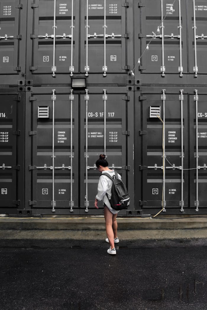 Woman with a backpack stands in front of gray shipping containers in an industrial area.